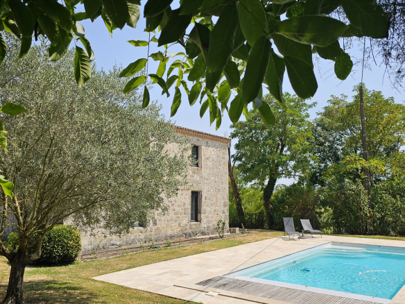 Stone cottage beside a rectangular swimming pool, surrounded by trees and a sunlit garden.