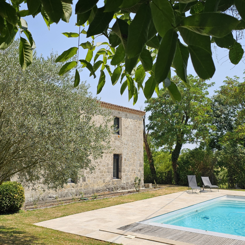 Stone cottage beside a rectangular swimming pool, surrounded by trees and a sunlit garden.