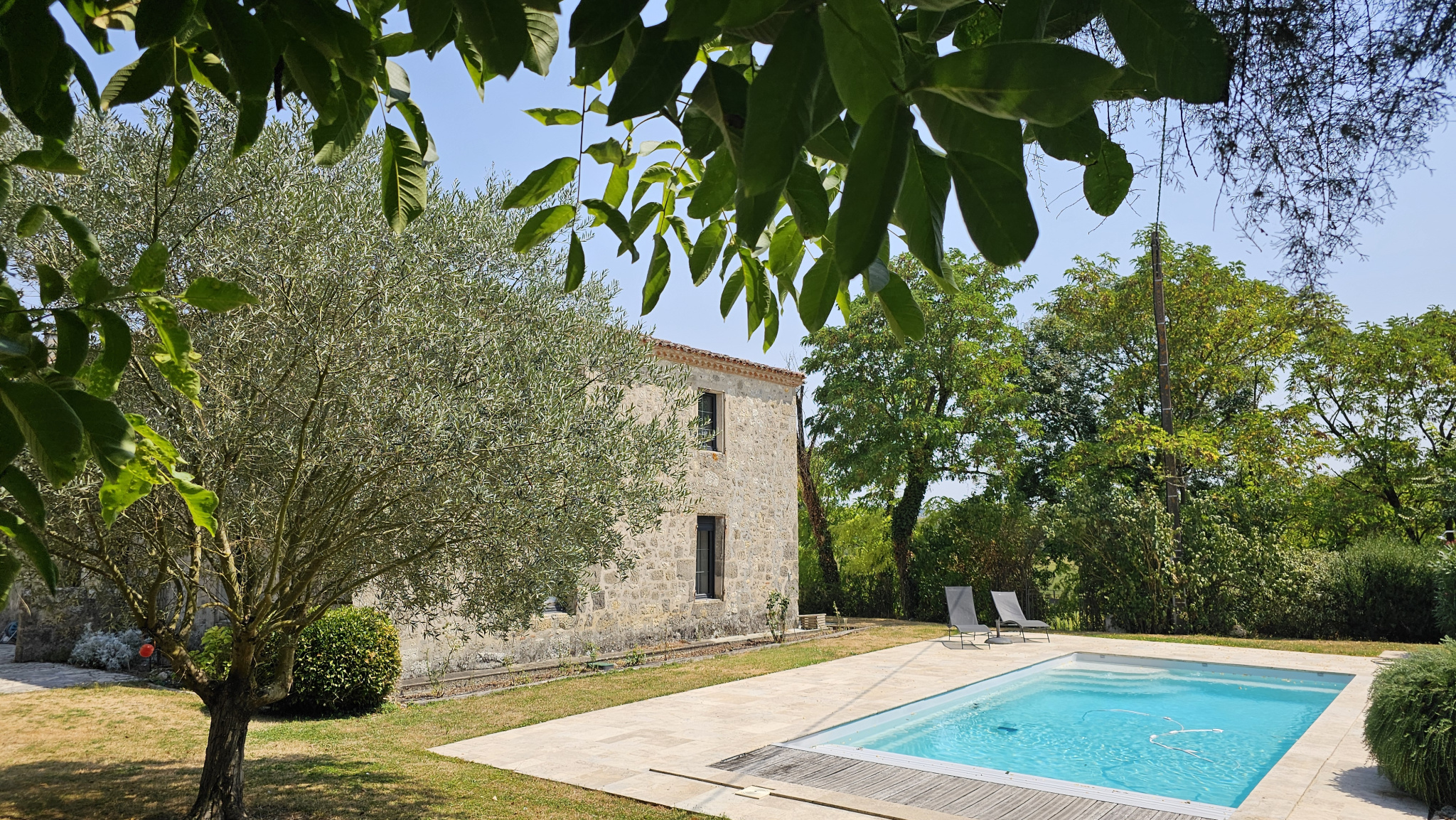 Stone cottage beside a rectangular swimming pool, surrounded by trees and a sunlit garden.