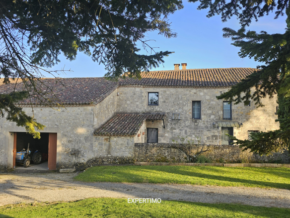 Stone farmhouse with a tiled roof, a small courtyard, and a blue tractor visible inside an open barn door, framed by overhead branches.