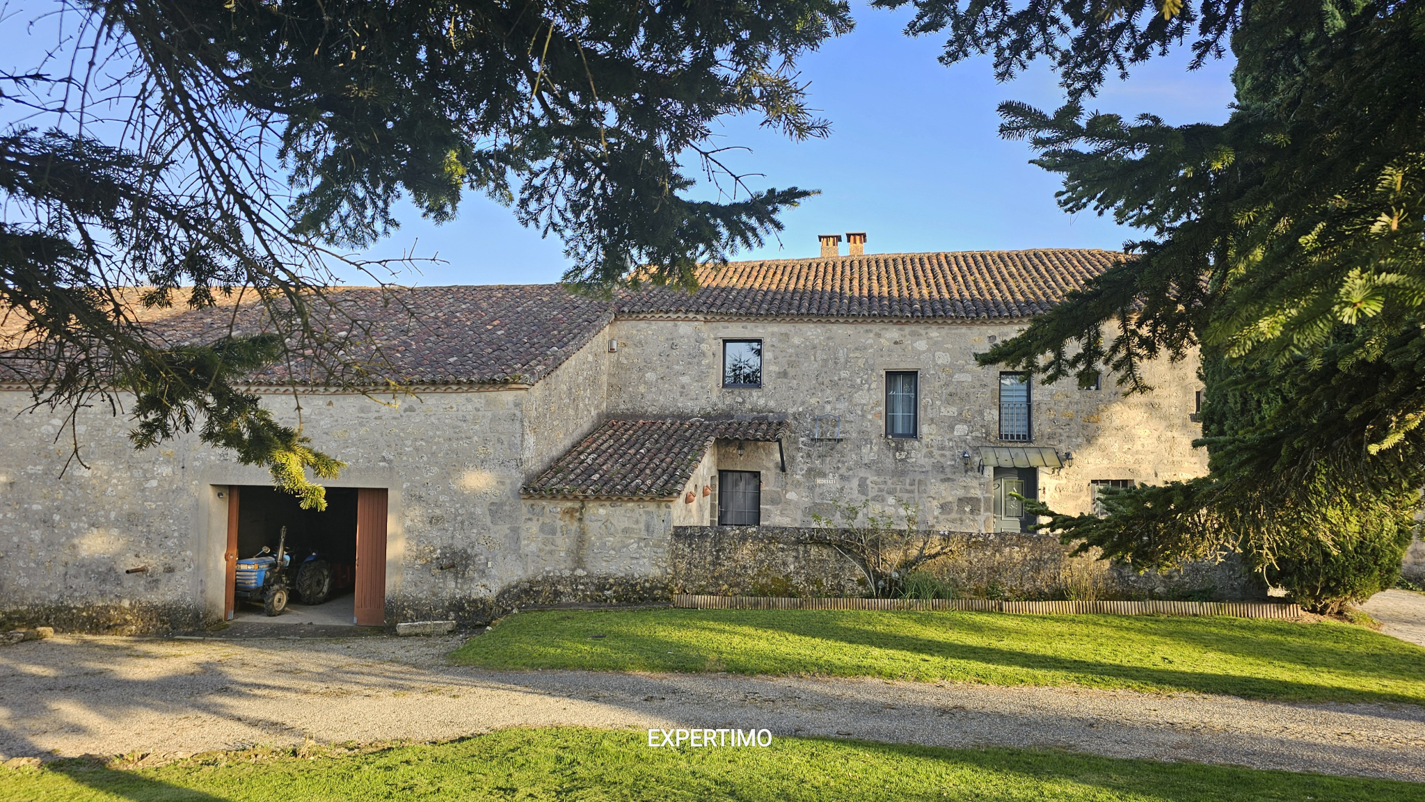 Stone farmhouse with a tiled roof, a small courtyard, and a blue tractor visible inside an open barn door, framed by overhead branches.