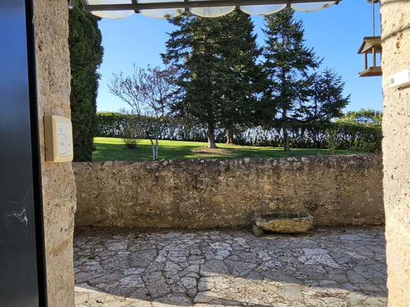 View from a stone doorway into a sunny garden with trees, a low stone wall, and a translucent glass canopy overhead.