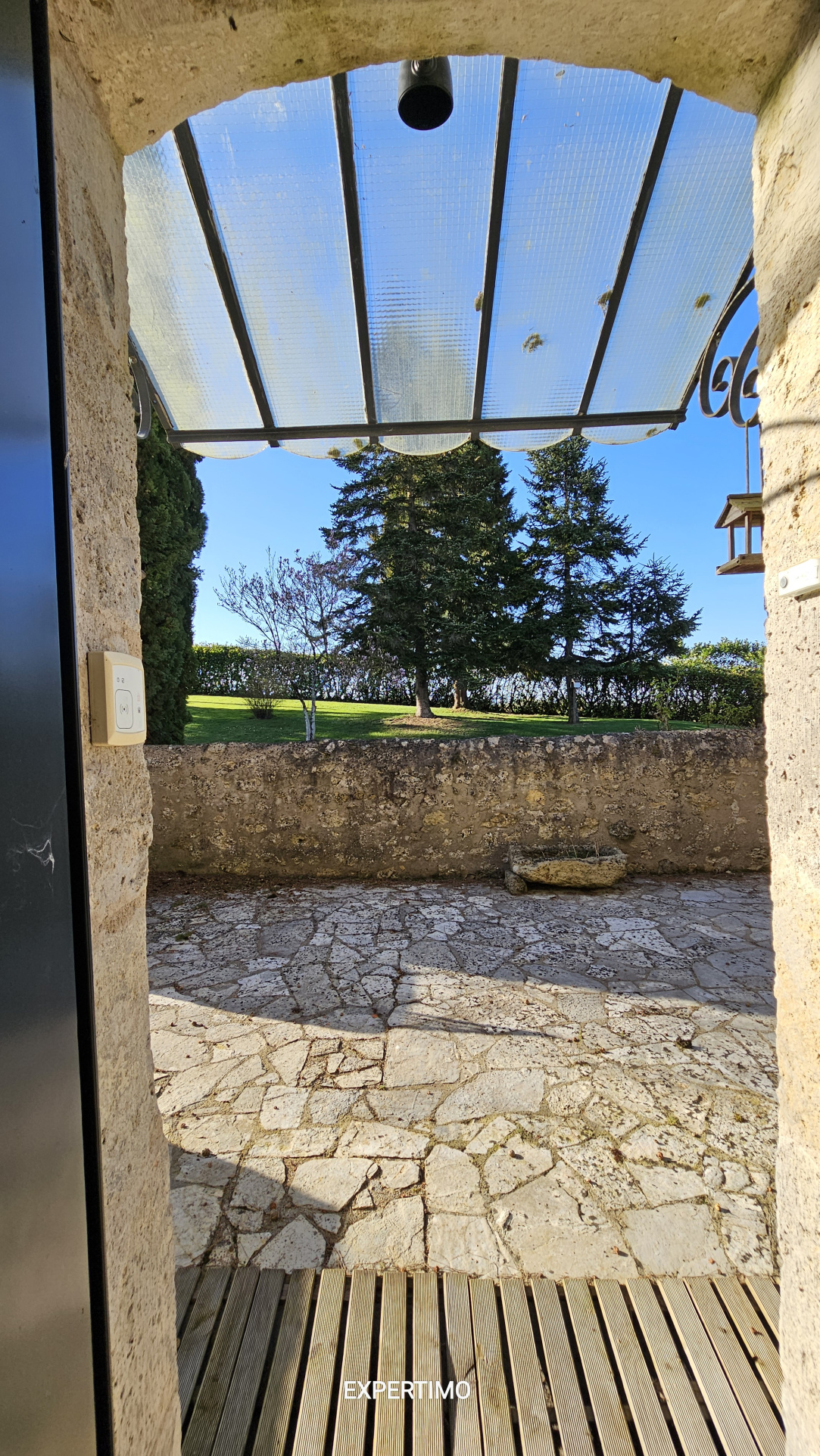 View from a stone doorway into a sunny garden with trees, a low stone wall, and a translucent glass canopy overhead.