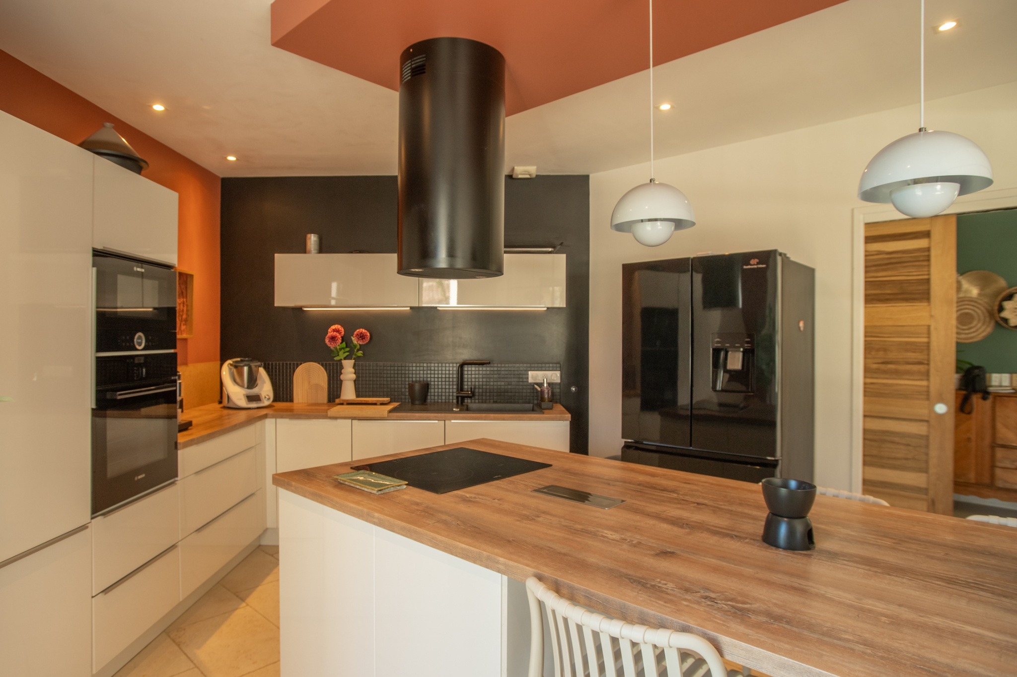 Modern kitchen with white cabinetry, wood island, black backsplash, and orange accent wall; pendant lights overhead.