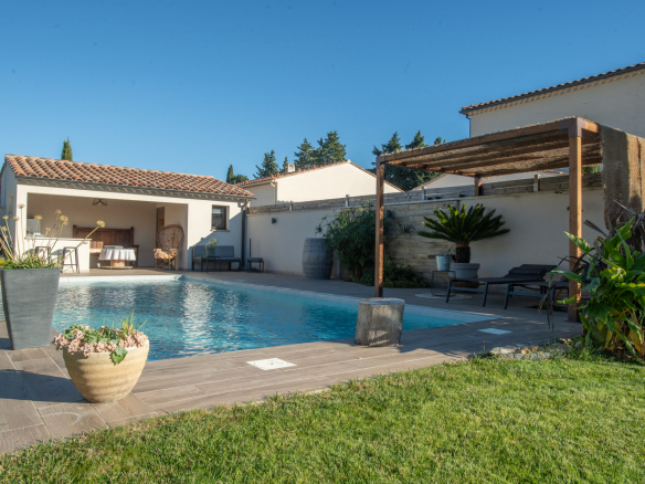 Backyard pool scene with a rectangular blue pool, beige tile deck, and a covered seating area on the left under a tiled roof.