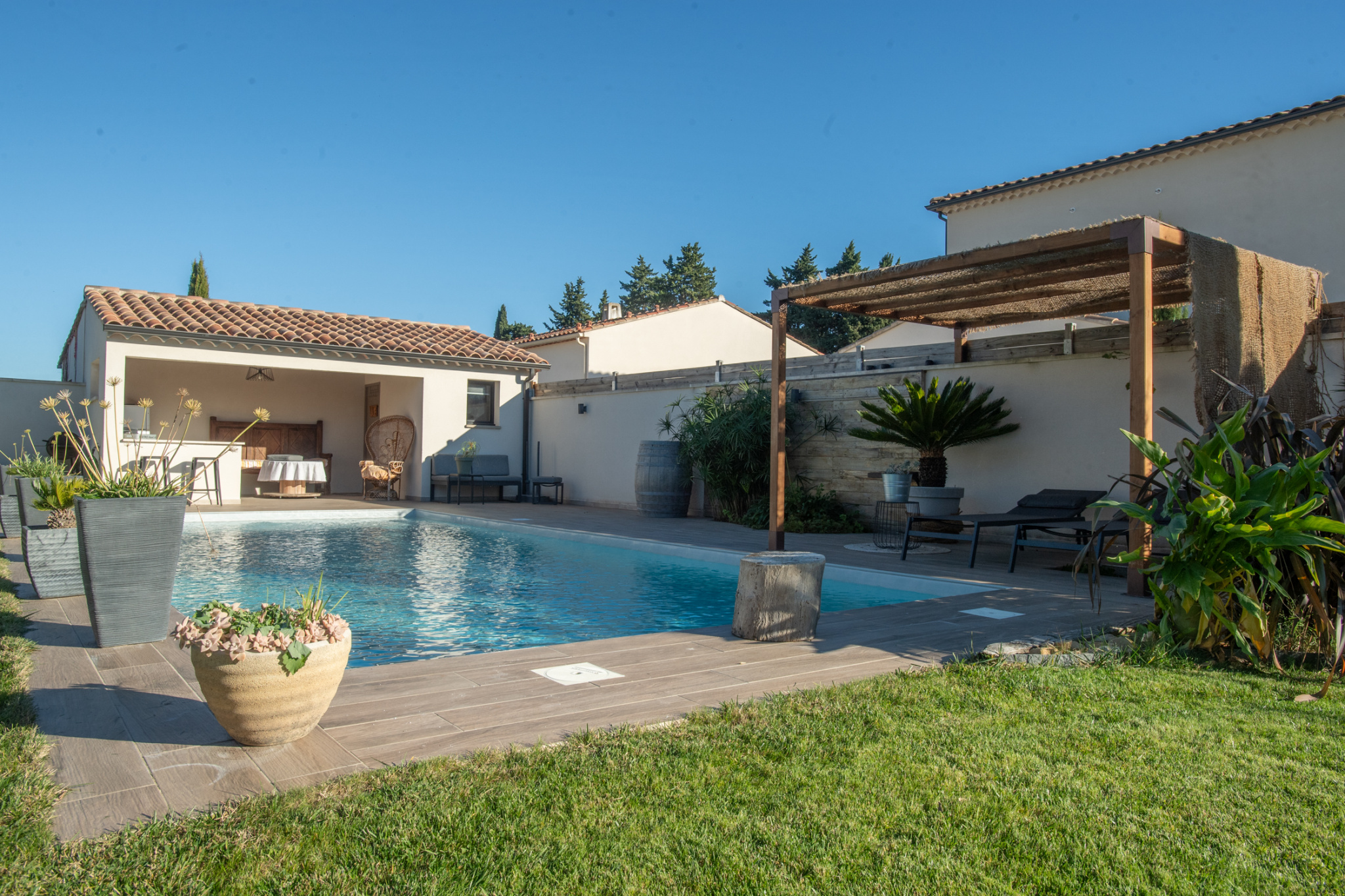 Backyard pool scene with a rectangular blue pool, beige tile deck, and a covered seating area on the left under a tiled roof.