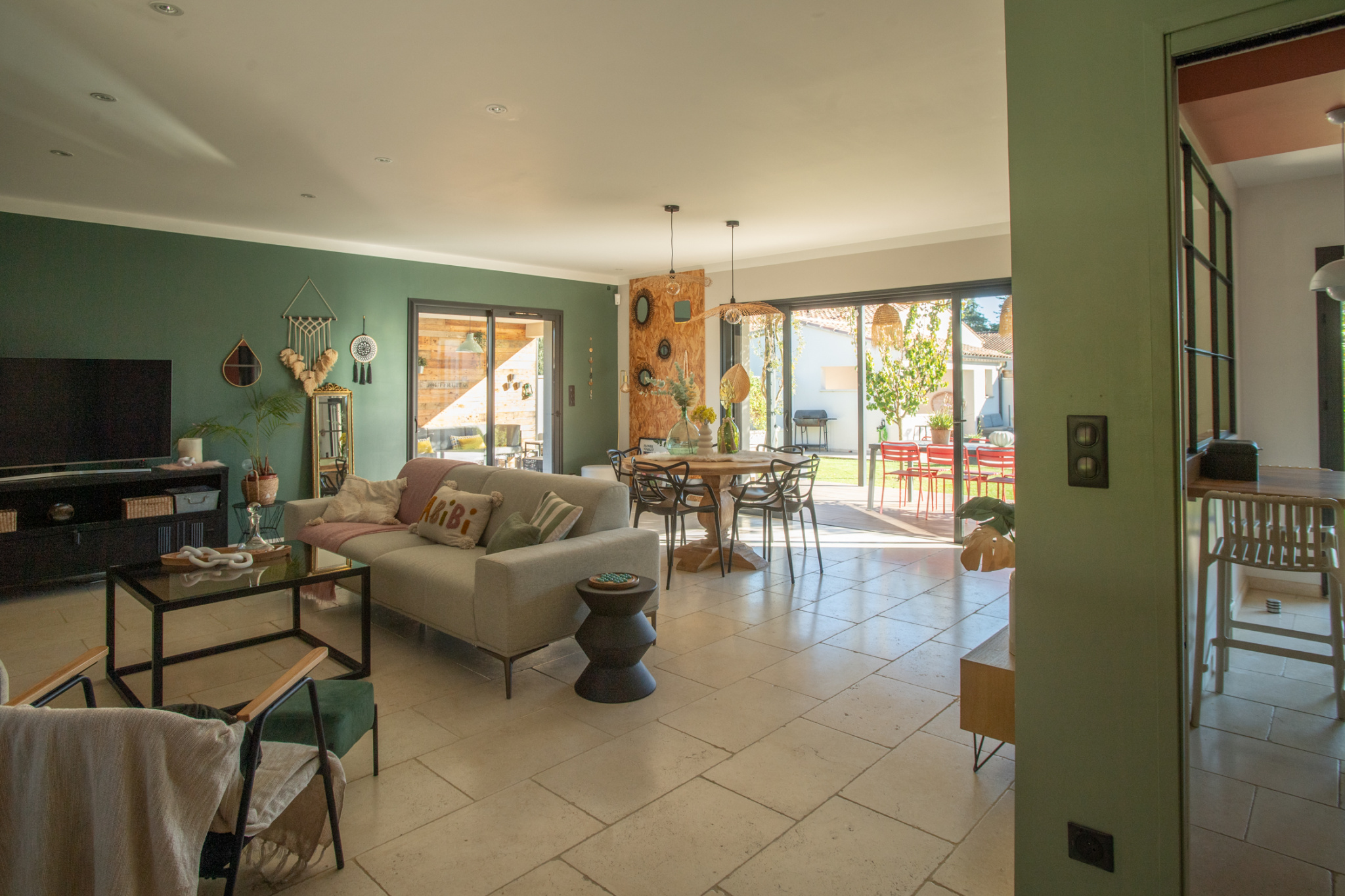 Open-plan living room with a green accent wall, beige sofa, TV cabinet, and a dining area near glass doors.