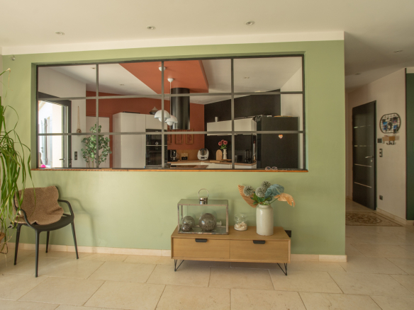 Open-plan living area with a green accent wall, a wooden console table with decor, and a glass-framed partition revealing the modern kitchen beyond.
