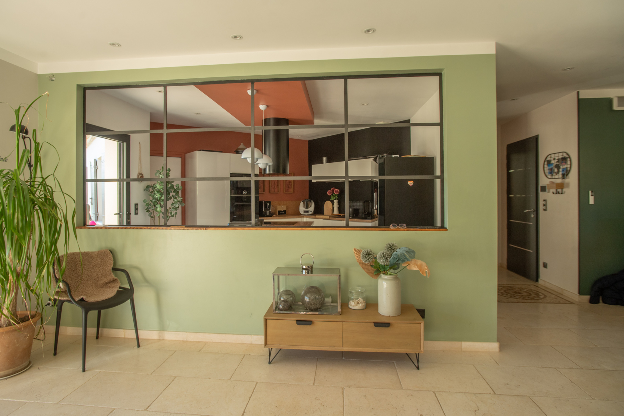 Open-plan living area with a green accent wall, a wooden console table with decor, and a glass-framed partition revealing the modern kitchen beyond.