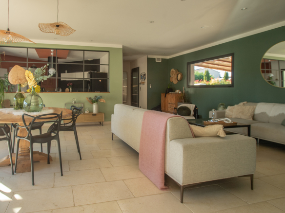 Open-plan living and dining room with green walls, round wooden table, black chairs, beige sofa, and plants.