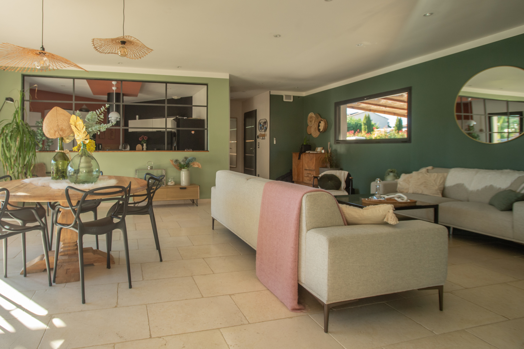 Open-plan living and dining room with green walls, round wooden table, black chairs, beige sofa, and plants.