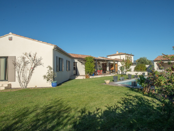 Backyard of a white villa with a red tile roof, green lawn, potted plants, and a pool beside a covered patio.