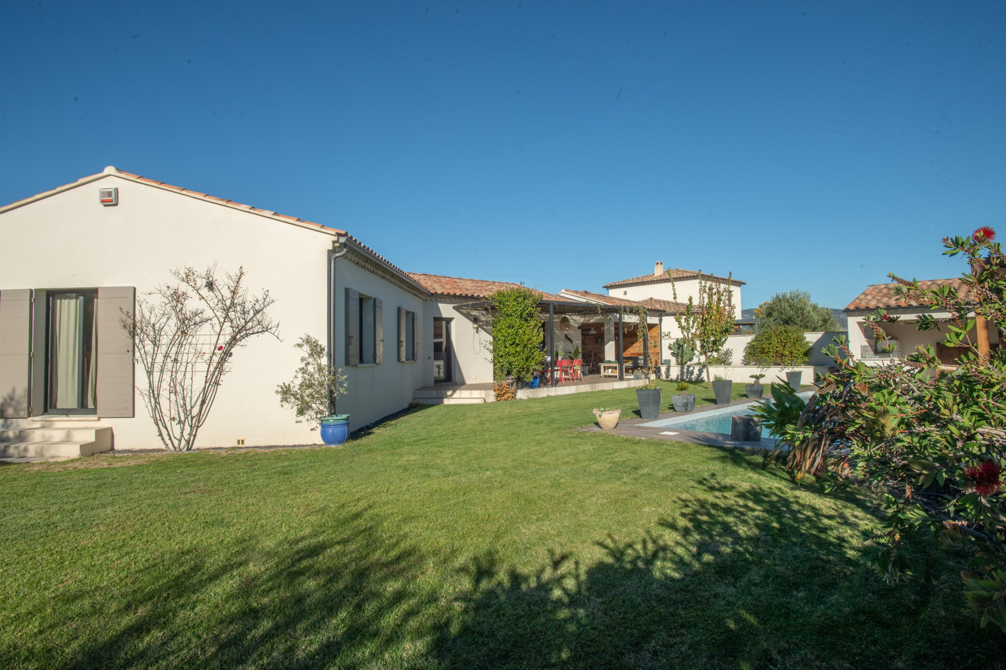 Backyard of a white villa with a red tile roof, green lawn, potted plants, and a pool beside a covered patio.