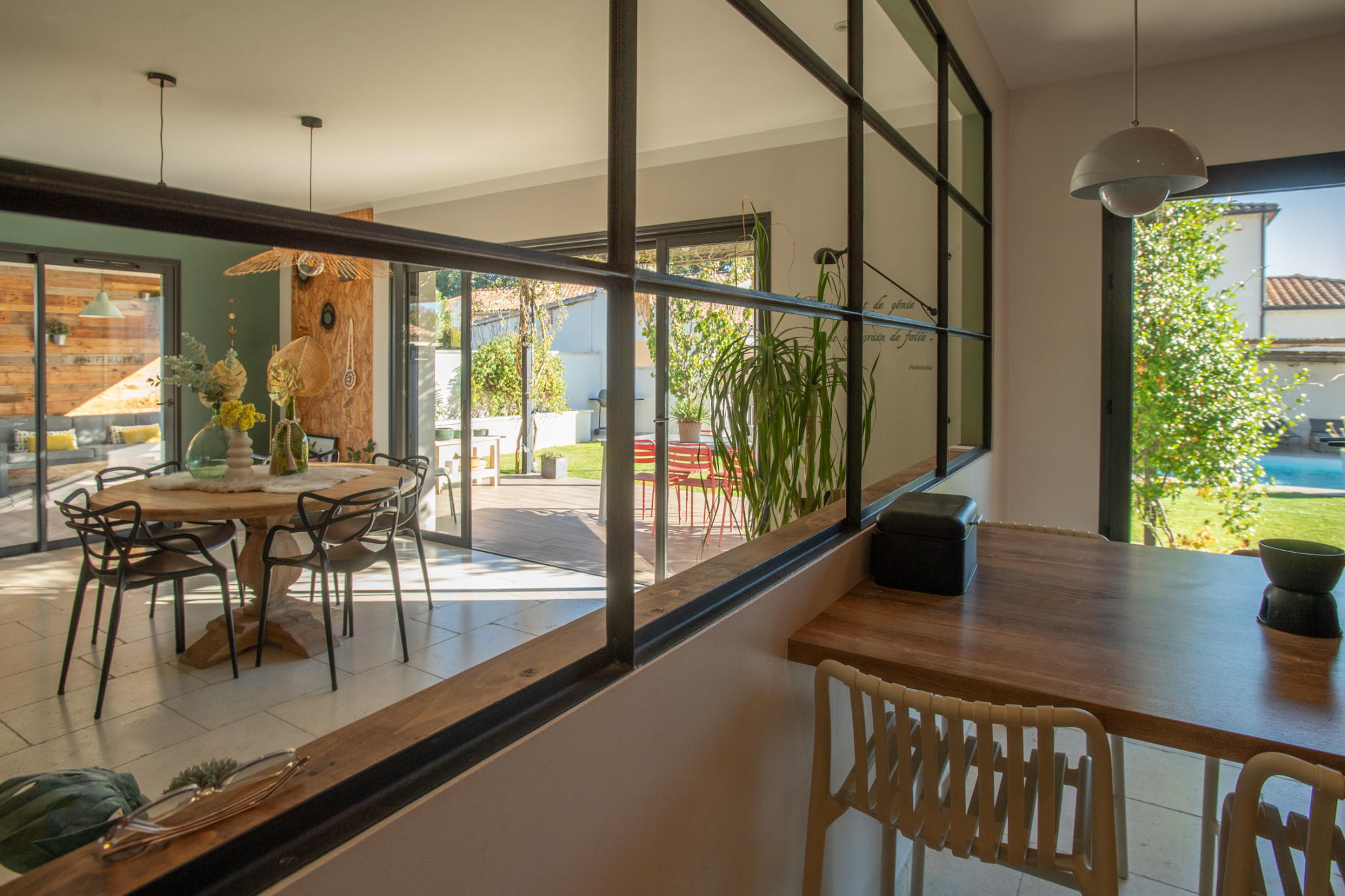 Round wooden dining table with black chairs in a sunlit open-plan dining area, separated from the outdoor patio by a glass partition.