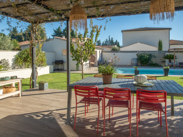 Patio dining area under a pergola with a gray table and red chairs, overlooking a rectangular pool in a sunny backyard.