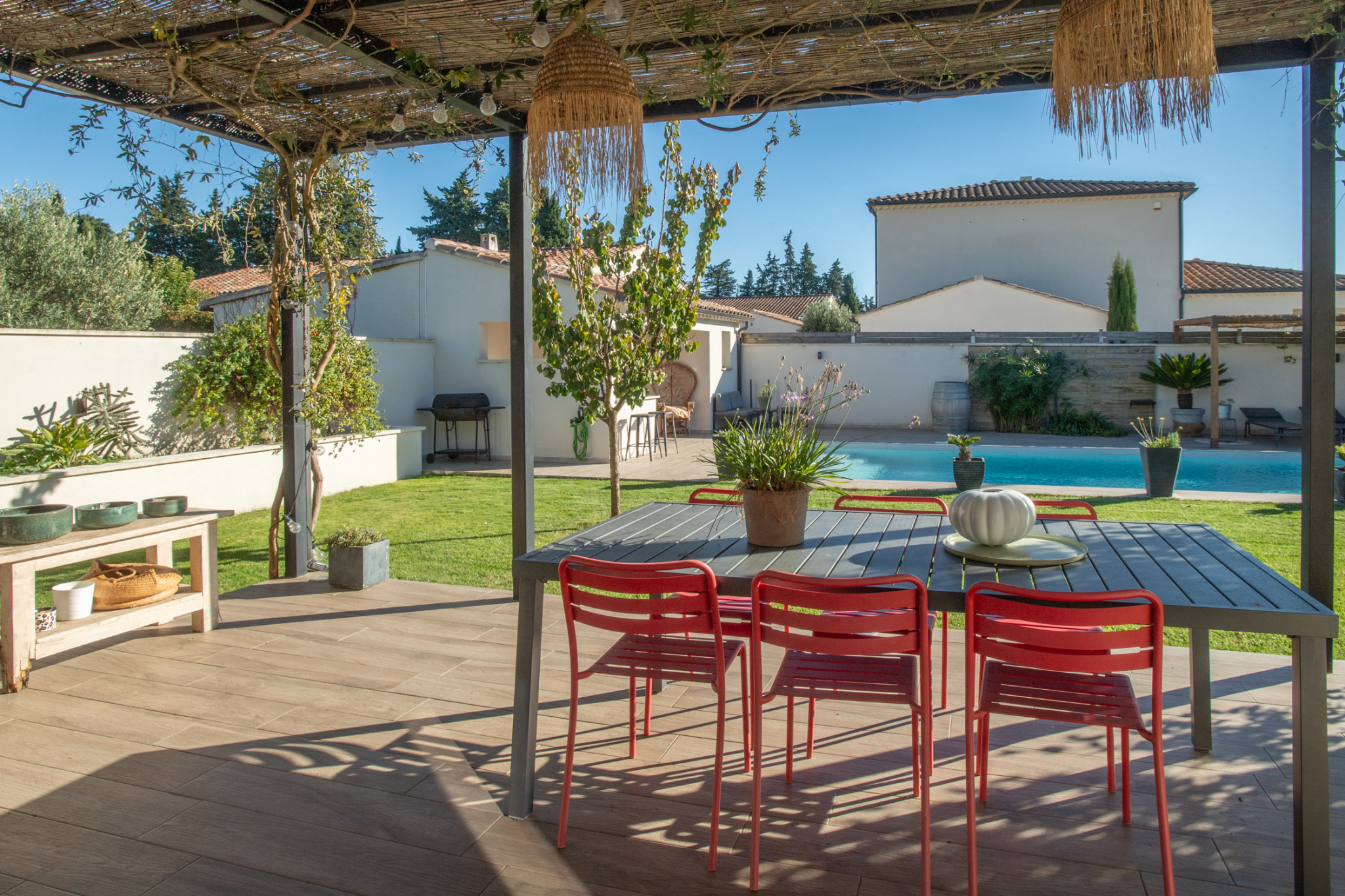 Patio dining area under a pergola with a gray table and red chairs, overlooking a rectangular pool in a sunny backyard.