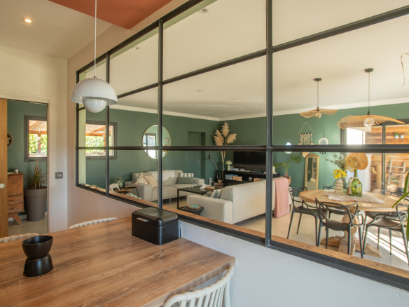 Open-plan living and dining area seen through a black-framed glass partition; wooden table in the foreground.