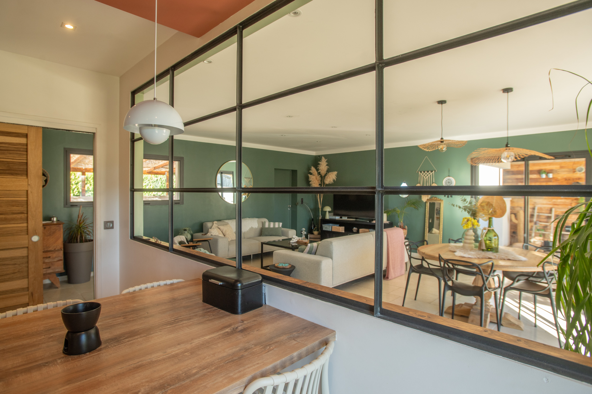 Open-plan living and dining area seen through a black-framed glass partition; wooden table in the foreground.