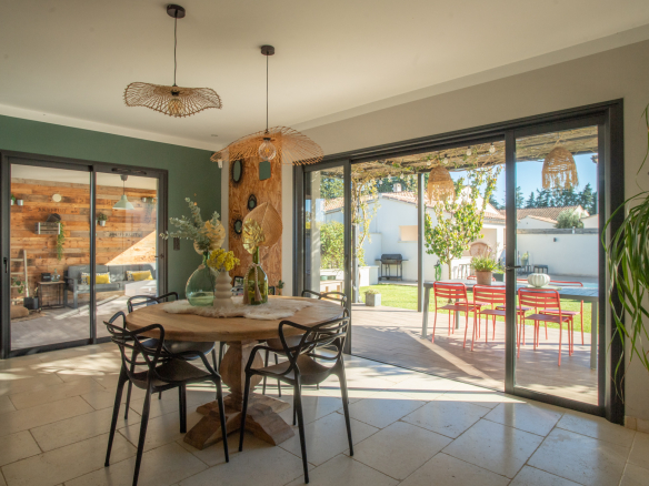 Sunny dining room with a round wooden table, black chairs, and woven pendant lights, opening to a patio with red chairs and greenery outside.
