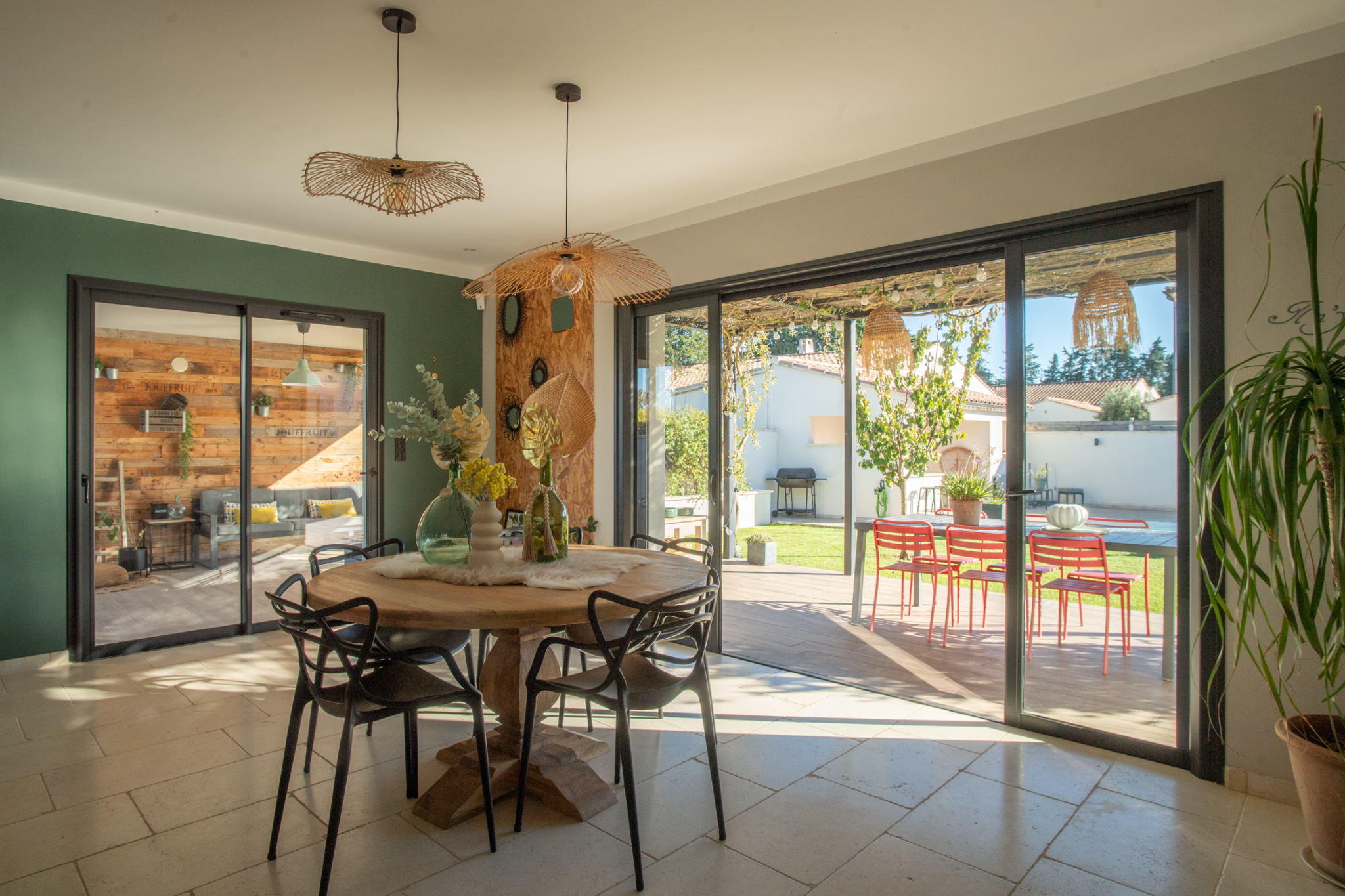 Sunny dining room with a round wooden table, black chairs, and woven pendant lights, opening to a patio with red chairs and greenery outside.