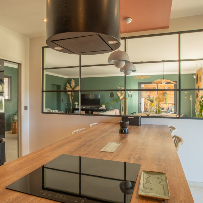 Long wooden kitchen island with an electric cooktop, black range hood above, and three metal pendant lights; glass-panel divider opens to a green-walled living area with plants and furniture.