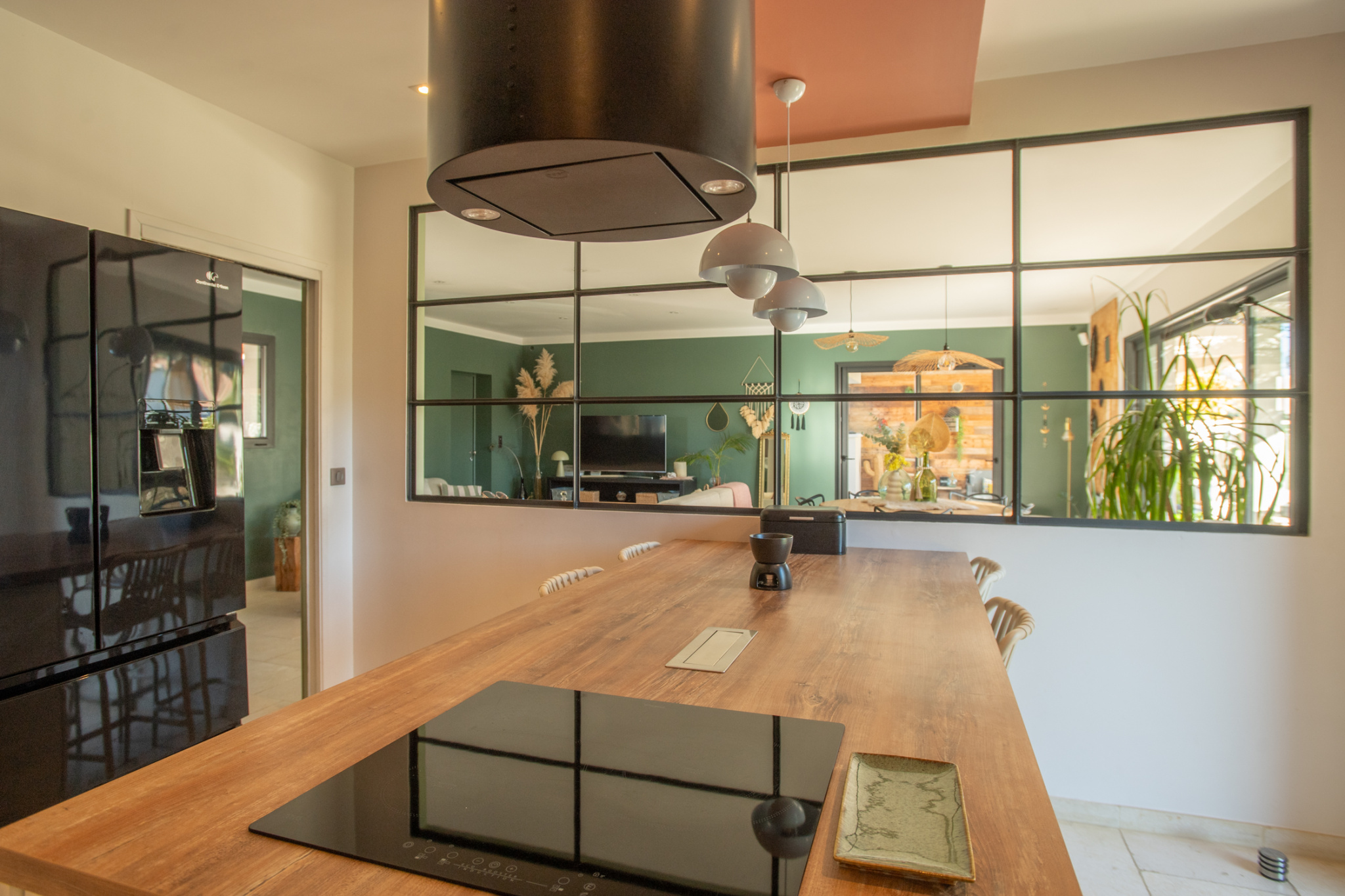 Long wooden kitchen island with an electric cooktop, black range hood above, and three metal pendant lights; glass-panel divider opens to a green-walled living area with plants and furniture.