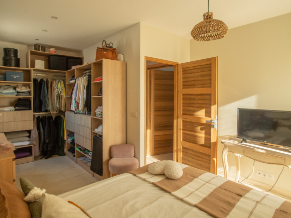 Bedroom with an open wooden wardrobe filled with clothes, shelves and boxes, next to a bed with a striped throw and pillows, TV on a white desk nearby.