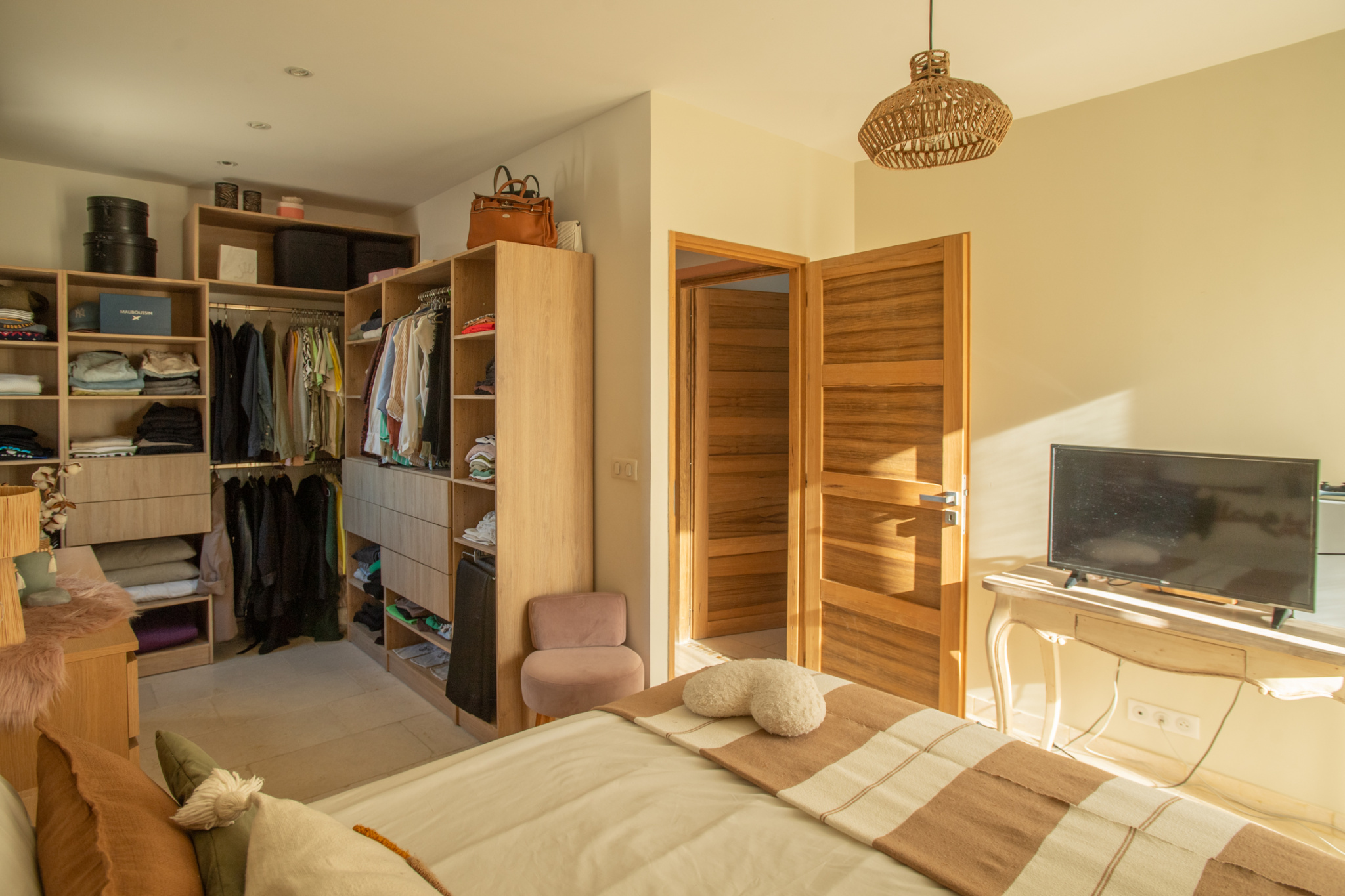 Bedroom with an open wooden wardrobe filled with clothes, shelves and boxes, next to a bed with a striped throw and pillows, TV on a white desk nearby.