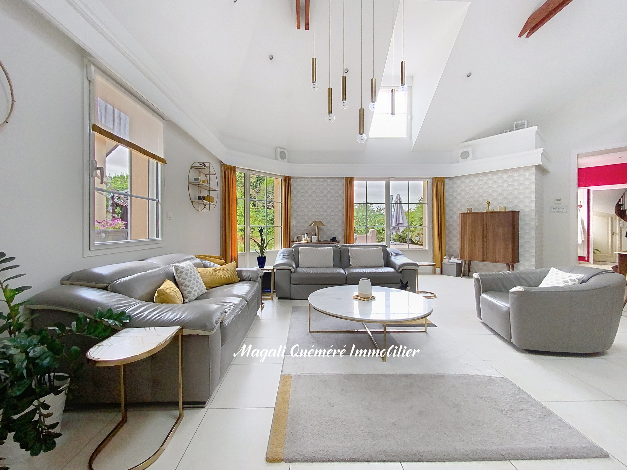 Bright, open living room with grey sectional sofas, a round marble coffee table, and mustard-yellow curtains by large windows letting in natural light.