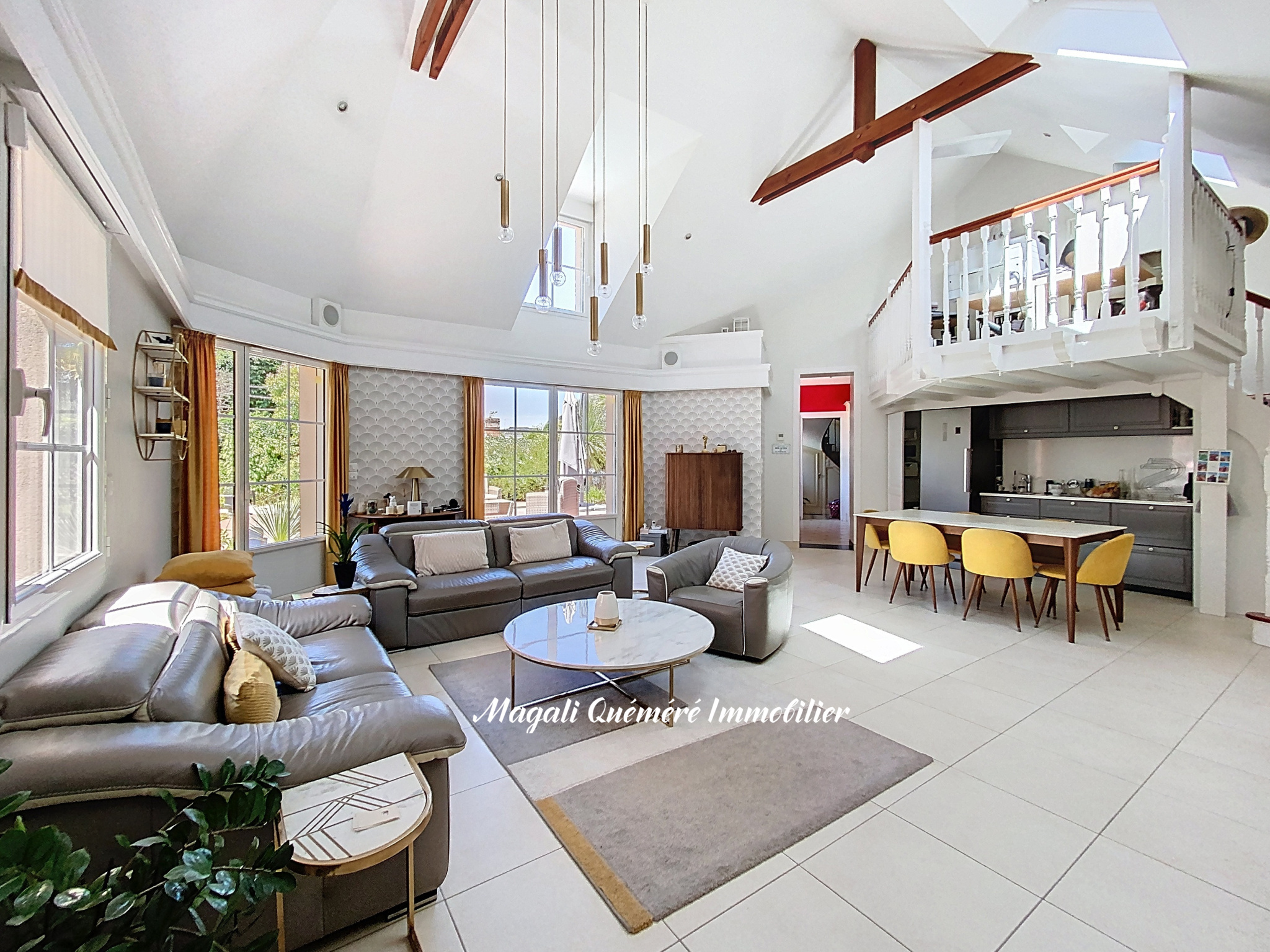Bright open-plan living area with grey leather sofas, round marble coffee table, and large windows to a garden. A white mezzanine loft overlooks a grey-kitchen/dining zone.