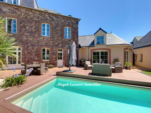 Backyard of a stone-brick house with a wooden deck, turquoise rectangular pool, and wicker seating under a clear blue sky.