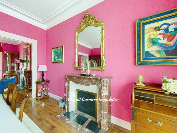 A bright pink living room with a marble fireplace, a large ornate gold mirror above it, and colorful abstract paintings on the wall; wooden floor and a sideboard with flowers on the right.