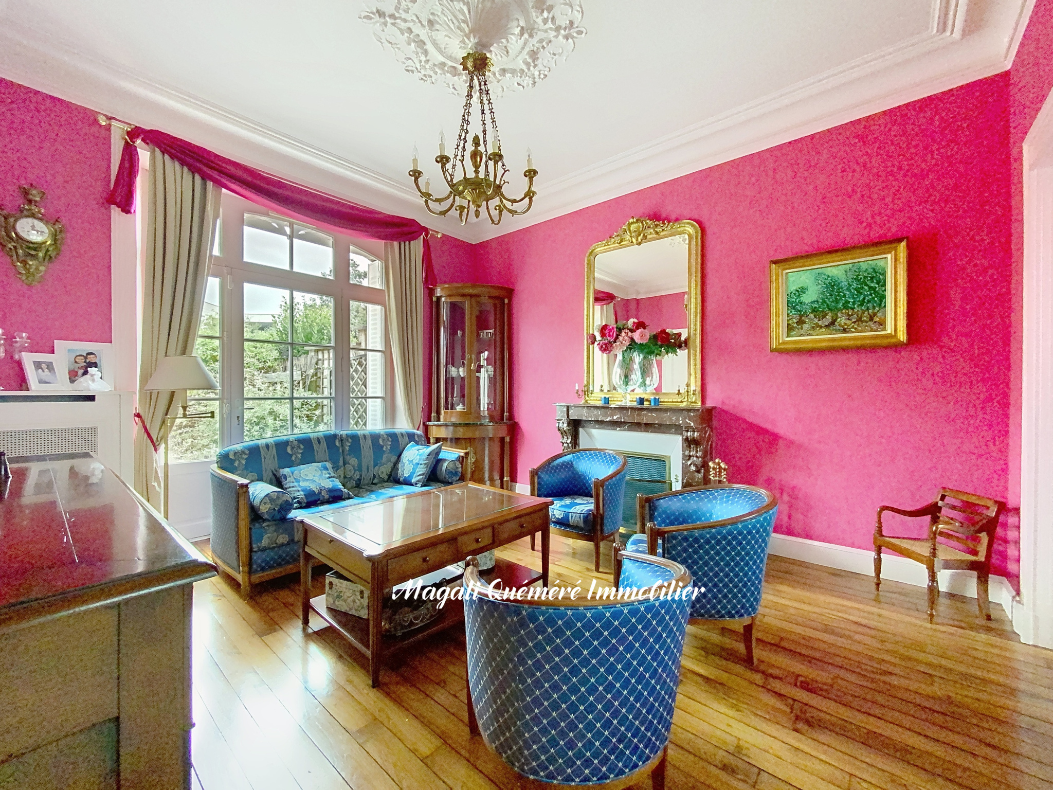 Bright pink living room with blue patterned furniture, a chandelier, and a marble fireplace with a gold-framed mirror.