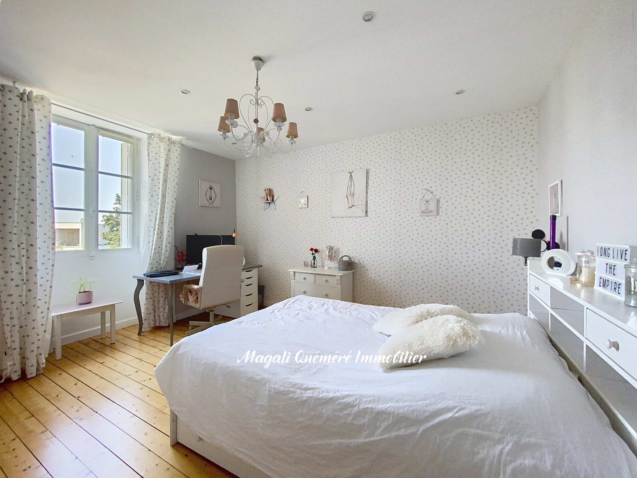 Bright bedroom with a white bed, chandelier, and large window with star-pattern curtains; pale walls and light wood floor.