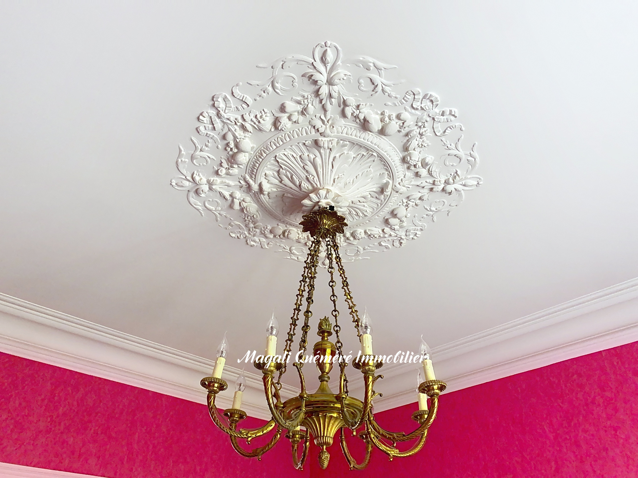White ornate ceiling medallion with floral scrollwork from which a gold chandelier hangs on brass chains above a pink wall interior.