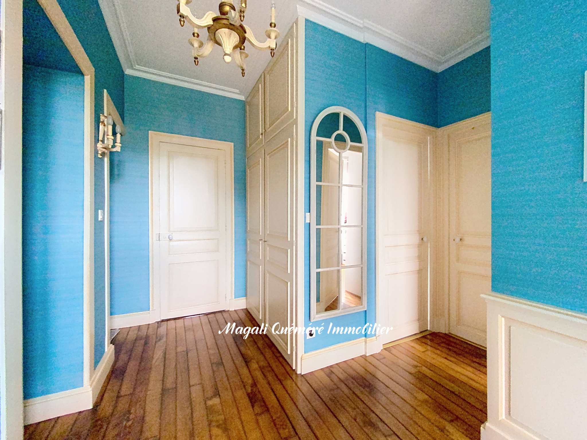 Blue-walled hallway with white panel doors, a tall built-in cabinet, and a wooden floor; chandelier overhead.