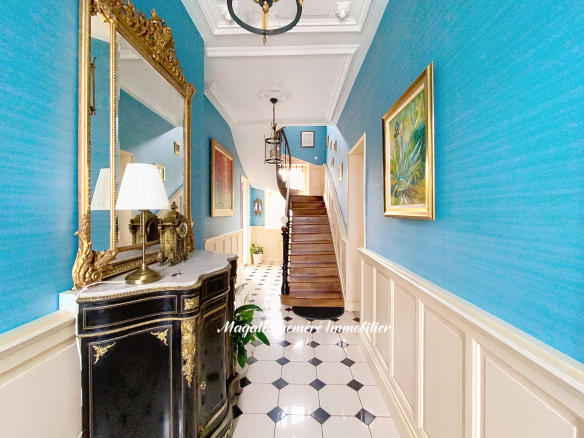 Elegant blue foyer with a black-and-gold antique console, gilded mirror, and chandelier along a checkered floor.