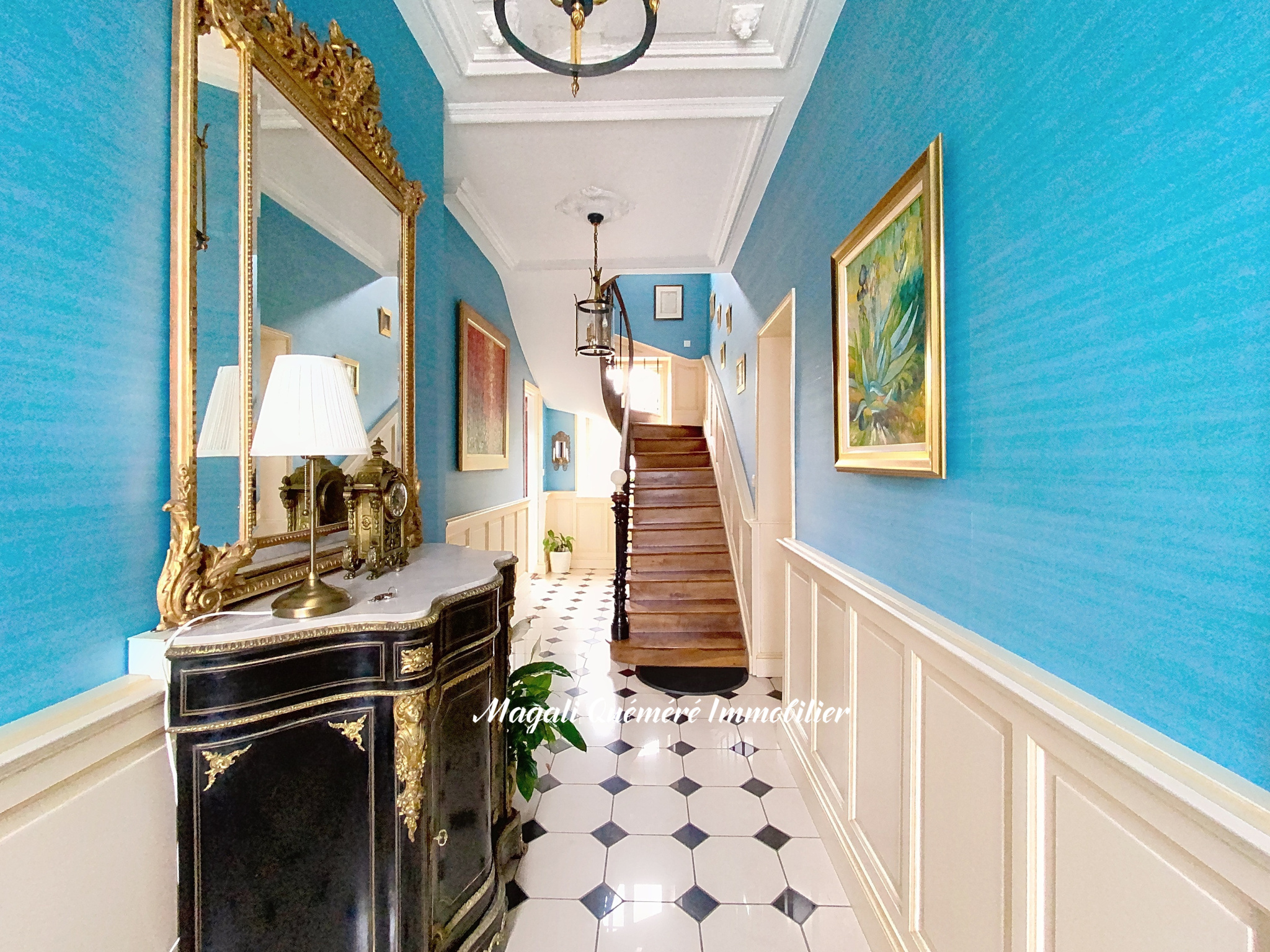 Elegant blue foyer with a black-and-gold antique console, gilded mirror, and chandelier along a checkered floor.