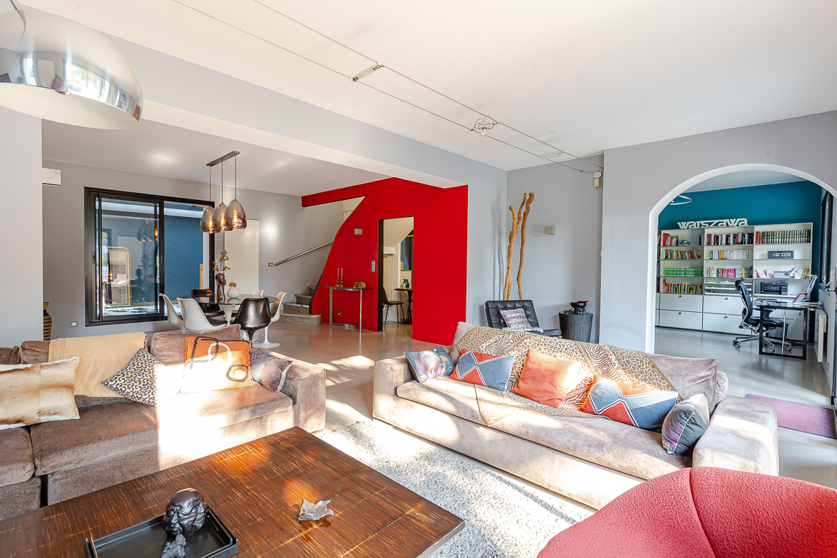 Spacious living room with beige sofas, colorful cushions, and a bold red geometric archway separating the space from the dining area.