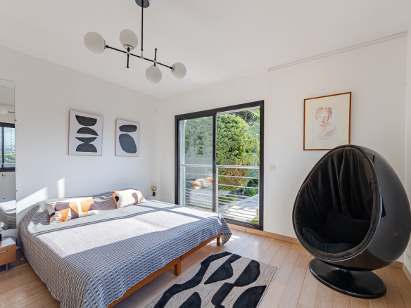 Bright modern bedroom with a low wooden bed, gray geometric bedspread, and a black egg-shaped lounge chair by a sliding balcony door.