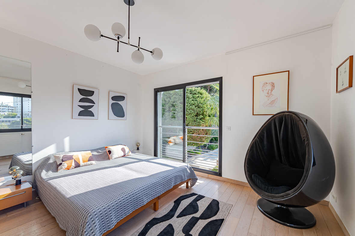 Bright modern bedroom with a low wooden bed, gray geometric bedspread, and a black egg-shaped lounge chair by a sliding balcony door.