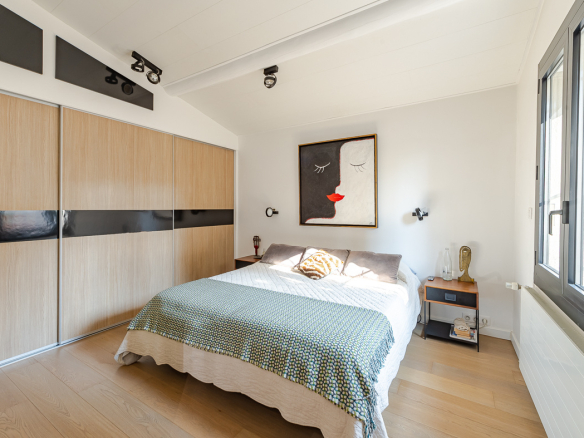 Modern bedroom with a large wooden wardrobe along the left wall, a centered bed, and a nightstand by the window under abstract art.