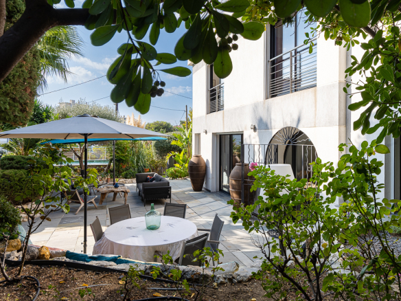 Modern backyard patio with a round table under a large umbrella, surrounded by lounge seating and lush greenery beside a white building.
