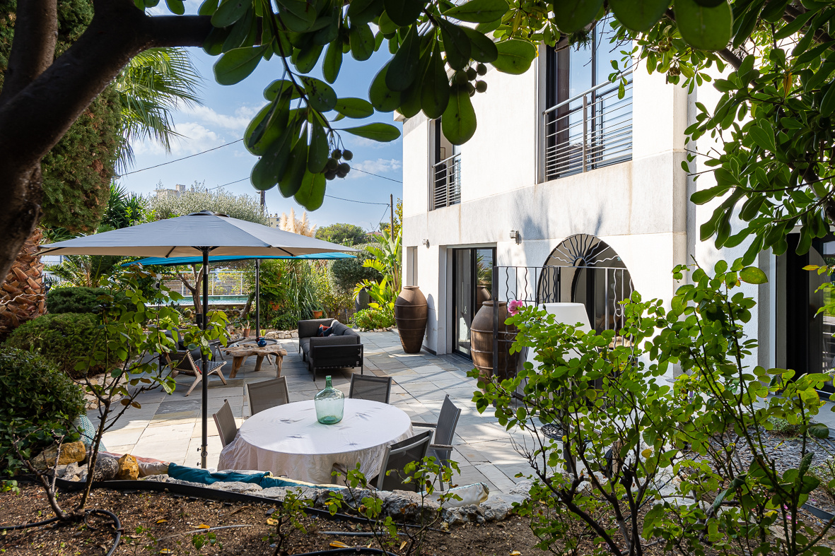 Modern backyard patio with a round table under a large umbrella, surrounded by lounge seating and lush greenery beside a white building.