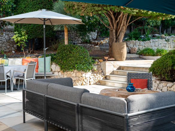 Outdoor patio with a gray sectional sofa, wooden coffee table, and blue vase under a large umbrella amid stone walls and greenery.