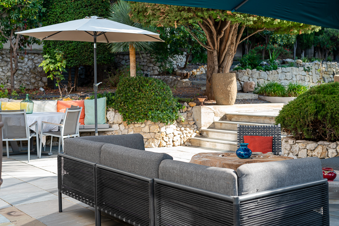 Outdoor patio with a gray sectional sofa, wooden coffee table, and blue vase under a large umbrella amid stone walls and greenery.
