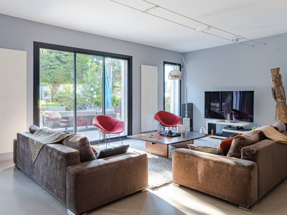 Bright modern living room with brown sofas, a wooden coffee table, and red accent chairs by large glass doors to the garden.