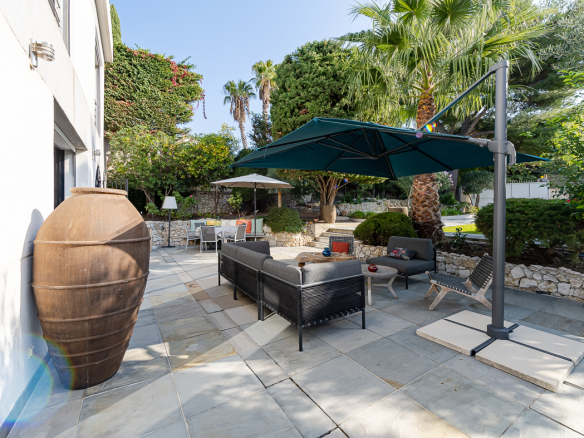 Outdoor patio with black wicker sofa set under a large blue umbrella, surrounded by palm trees and stone walls.
