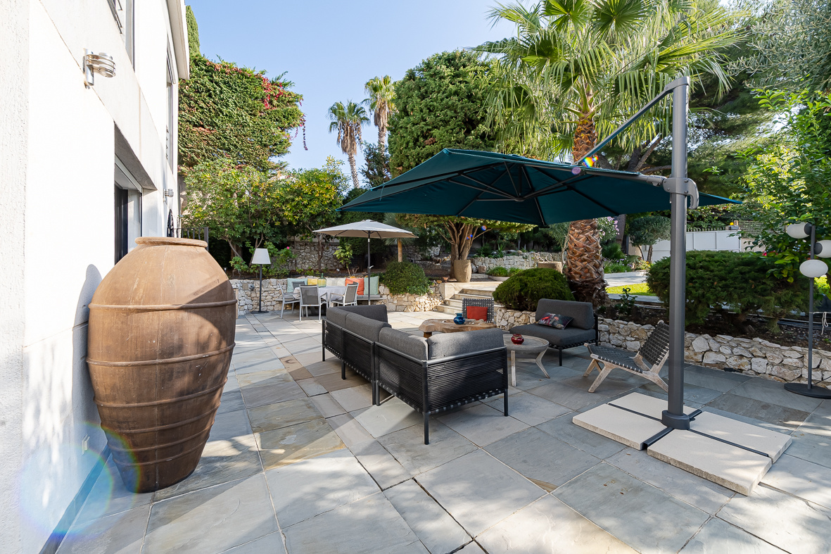 Outdoor patio with black wicker sofa set under a large blue umbrella, surrounded by palm trees and stone walls.