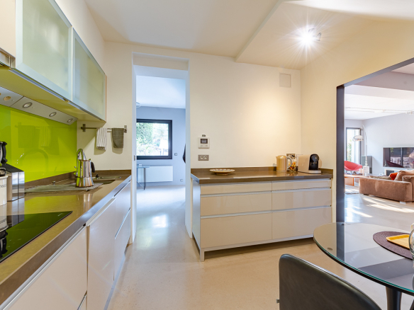 Modern kitchen with white cabinets, lime green backsplash, and stainless countertops; dining table and living room visible through an open doorway.